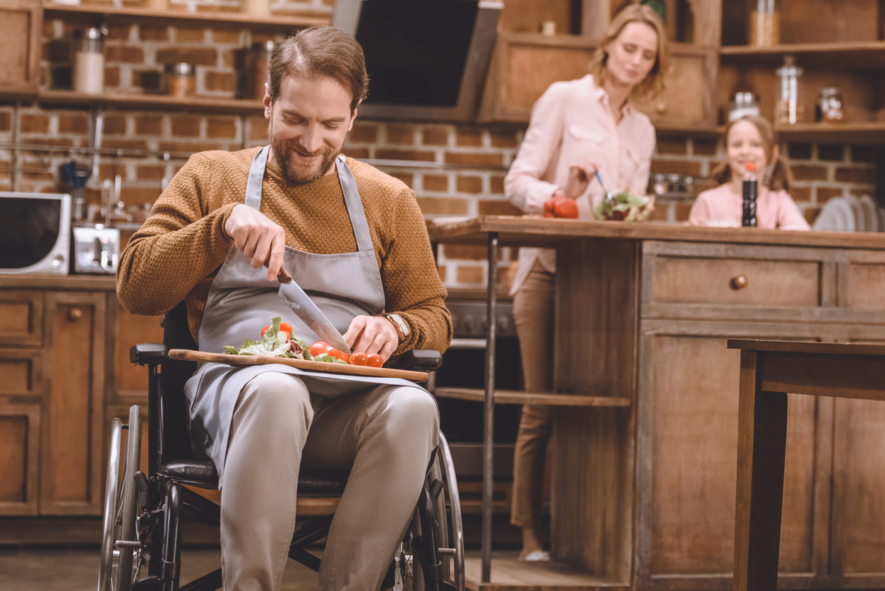 smiling man in wheelchair cutting vegetables for salad while happy mother and daughter standing behind